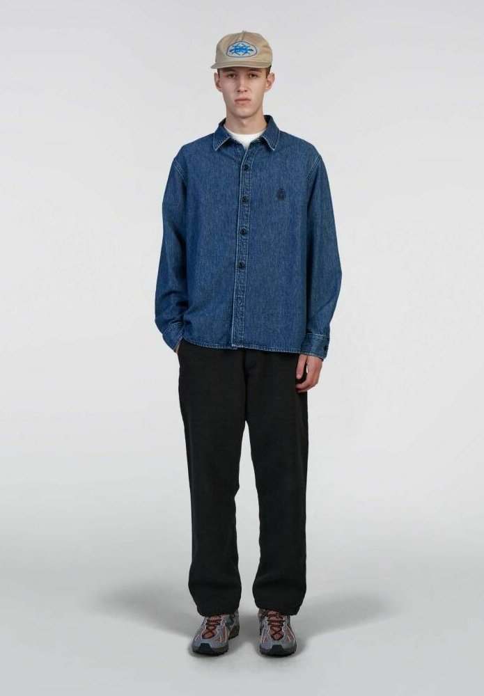 Camicia in denim con bottoni e colletto, abbinata a pantaloni scuri e sneakers. Cappello con logo circolare blu su sfondo beige.