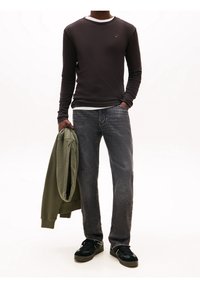 Man wearing black long-sleeve shirt, dark gray jeans, and black sneakers holding an olive green jacket in one hand.