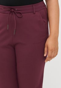Detailní záběr ruky osoby sahající k pase vínových kalhot se šňůrkou a postranními kapsami na bílém pozadí.