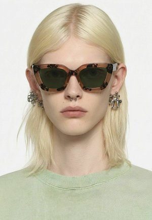 Personne aux cheveux blonds raides portant des lunettes de soleil écaille de tortue, un piercing au septum, des boucles d'oreilles en argent distinctives et un haut vert clair à col rond.