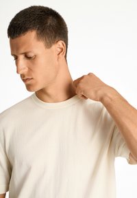 Krämfärgad t-shirt med kort ärm, ribbad textur, rund halsringning, figurnära silhuett, bärs av en person med kort hår som ser ner.