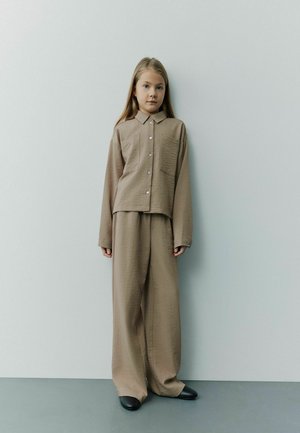 Beige strukturerad tvådelad outfit bestående av en kragklädd skjorta med knappar och fickor samt vida byxor, tillsammans med svarta skor.