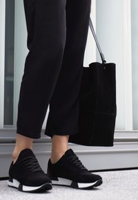 Černé sportovní boty s texturou síťoviny a semiše mají bílou mezipodešev. V jedné ruce je držena černá semišová taška, která je zkombinována s černými kalhotami.