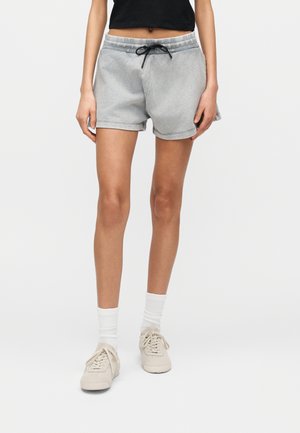 Osoba oblečená v sivých šortkách so šnúrkou, biele ponožky, béžové tenisky a čierny krátky top na jednoduchom svetlom pozadí.