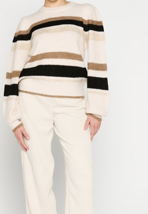 Personne portant un pull crème avec des rayures horizontales noires et marron et un pantalon crème à jambes larges, sur fond blanc uni.