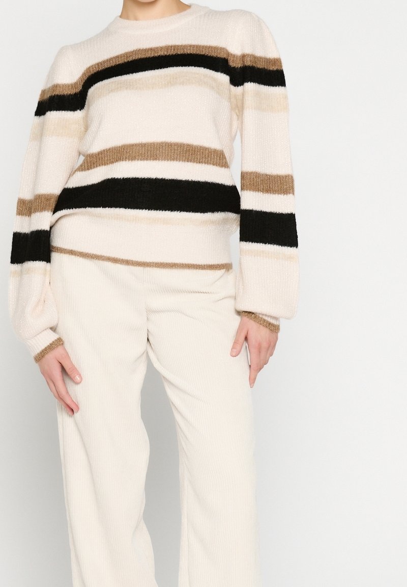 Personne portant un pull crème avec des rayures horizontales noires et marron et un pantalon crème à jambes larges, sur fond blanc uni.