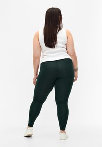 Groene legging gemaakt van rekbaar materiaal, met een strakke pasvorm en hoge taille, gecombineerd met een witte mouwloze top en witte sneakers.