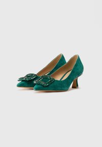 Décolleté in suede verde smeraldo con tacco basso, punta affusolata e grandi fibbie decorative con gioielli verdi su uno sfondo bianco semplice.