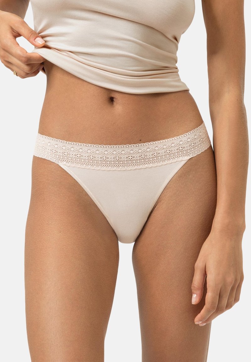 Frau hebt beiges Oberteil an, um passende beige Unterwäsche mit Spitzenbesatz vor einem schlichten, hellen Hintergrund zu zeigen.