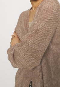 Ljusbrun stickad cardigan med lös passform, V-ringning och långa ärmar. Har en mjuk textur och synliga stickmönster.