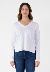 Maglione bianco a V con maniche lunghe, orlo a coste e texture morbida. Presenta un piccolo logo sul lato sinistro.
