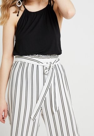 Vrouw draagt een mouwloos zwart topje en witte broek met hoge taille, verticale zwarte strepen en een gestrikte riem in de taille.