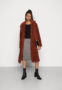 Manteau en fausse fourrure marron avec un col à revers, pull en tricot noir, jupe midi à carreaux, et bottines à talons noires. Petit sac à main noir inclus.