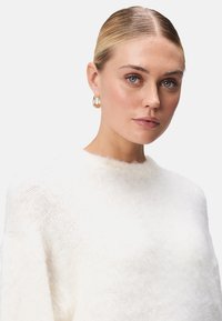 Biely, nadýchaný sveter s okrúhlym výstrihom; jemná textúra; modelka má na sebe zlaté kruhové náušnice; neutrálny pozadie, ktoré zvýrazňuje detaily oblečenia.