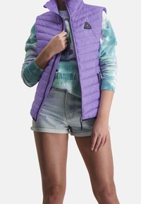 Fialová bezrukávová bunda s prošívaným designem, zapínání na zip a logem. Nositá přes tričko s barevným vzorem a světlé džíny šortky.