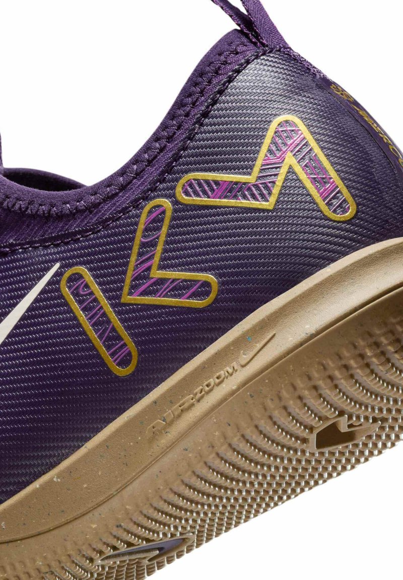 Zapatilla deportiva morada con parte superior sintética texturada, acentos en dorado y rosa, y suela de goma beige que presenta un patrón de tracción.