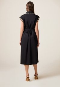 Robe noire à col avec manches courtes, taille élastique et longueur midi fluide. Détails en dentelle sur le col. Sandales à talons visibles.