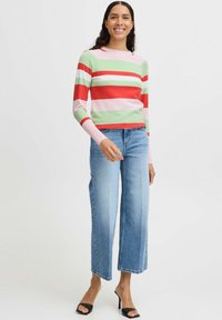 Pull en tricot rayé vert, rose, rouge et blanc, associé à un jean large bleu clair. Le mannequin porte des talons ouverts noirs.
