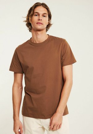 Junger Mann mit schulterlangem Haar, der ein schlichtes braunes T-Shirt mit kurzen Ärmeln und helle Hosen trägt, vor einem neutralen Hintergrund stehend.