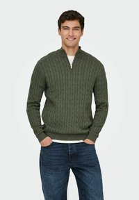 Pull en tricot vert avec un col haut et une fermeture �éclair, présentant un motif en relief. Assorti à un jean en denim bleu.