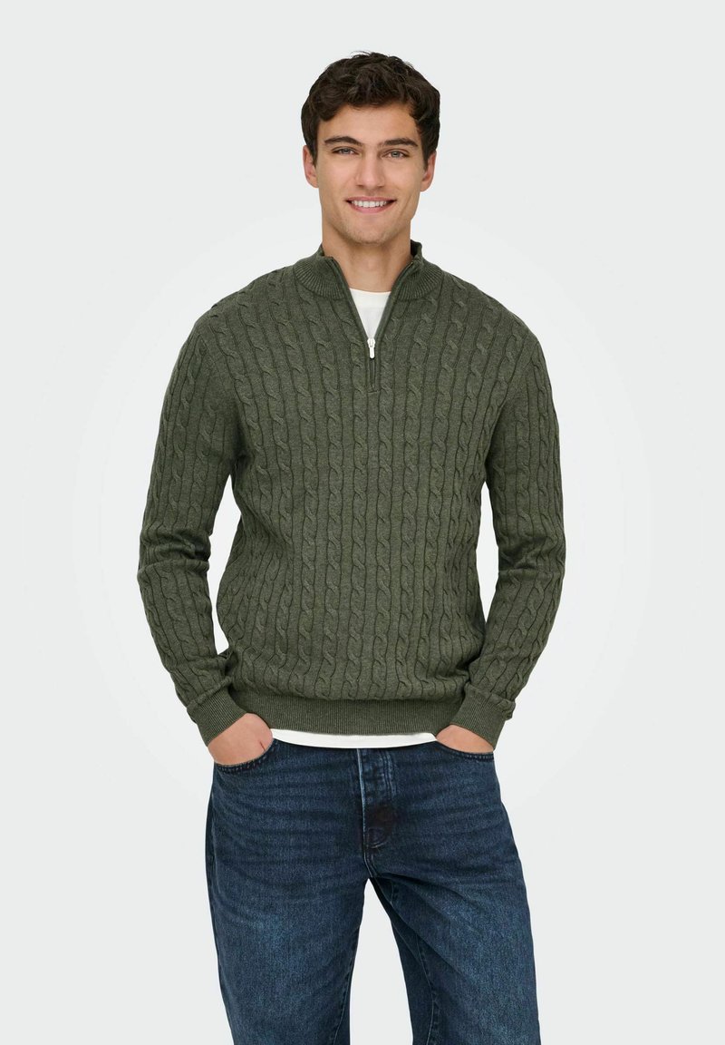 Pull en tricot vert avec un col haut et une fermeture éclair, présentant un motif en relief. Assorti à un jean en denim bleu.