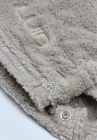 Manta de forro polar en beige claro con una superficie suave y texturizada, que cuenta con cierres a presión y un logotipo sutil bordado a lo largo del borde.