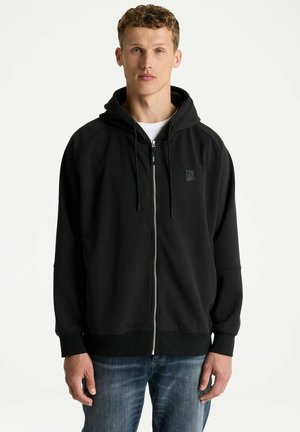 Junger Mann mit kurzen lockigen Haaren, der einen schwarzen Zip-Hoodie und blaue Jeans trägt, steht vor einem schlichten hellen Hintergrund.