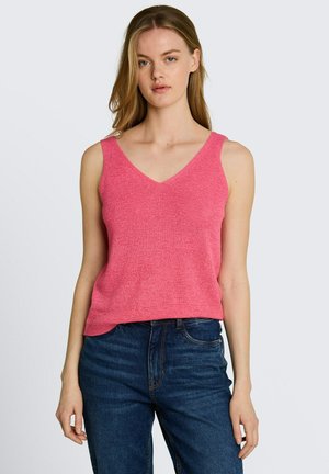 Frau trägt ein ärmelloses pinkes Stricktop mit V-Ausschnitt und blauen Denim-Jeans, steht vor einem schlichten hellen Hintergrund.