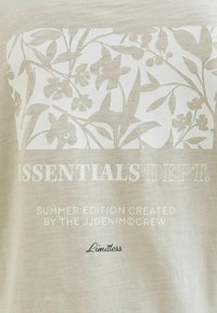 Lichtbeige t-shirt met een textuur bloemenprint in lichtere tinten, voorzien van de tekst "ESSENTIALS DEPT." en "Limitless" in contrasterende kleuren.