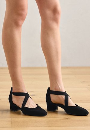 Stopy w czarnych butach na niskim obcasie z krzyżującymi się paskami przy kostce, na jasnej drewnianej podłodze na tle jednolitego tła.