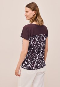 Kortærmet top med en maroon stribet ryg og hvid front, der har et dristigt, mørkt bladmønster. Blødt stof, afslappet fit.