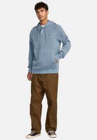 Light blue hooded sweatshirt with a textured fabric, front pocket, and drawstrings, paired with brown pants and black sneakers.