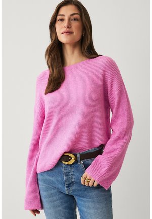 Kvinde med langt brunt hår iført en pink ribstrikket sweater, blå jeans, sort bælte med guldlås og gyldne ringe på højre hånd.