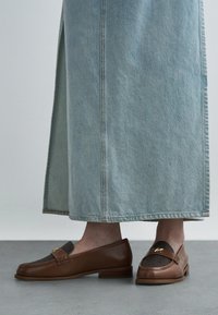 Mocassins de couro castanho com painel de tecido cinzento, detalhe em dourado, combinados com uma saia de ganga azul claro, mostrando a biqueira arredondada do sapato.