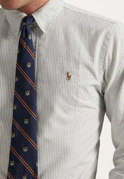 Pruhovaná šedá košeľa s malým vyšitým logom polo a námorníckou kravatou s diagonálnymi pruhmi a opakujúcimi sa erbovými symbolmi.