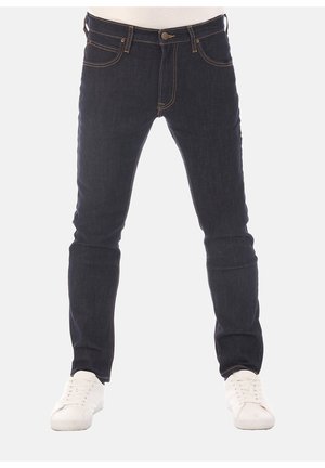 Jeans aderenti blu scuro indossati con sneakers bianche, mostrati dalla vita ai piedi su uno sfondo chiaro e semplice.