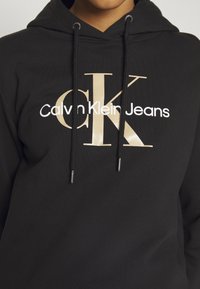 Černá mikina s kapucí a šňůrkami, na přední straně s velkým zlatým logem "CK" a bílým textem "Calvin Klein Jeans".