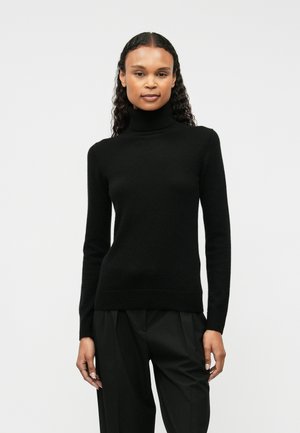 Maglione nero a collo alto in materiale morbido, design aderente con maniche lunghe e orlo a costine, indossato con pantaloni neri larghi.