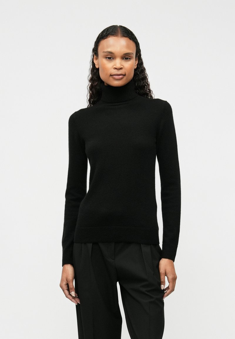 Fekete garbós pulóver puha anyagból, testhezálló fazon, hosszú ujjakkal és bordás szegéllyel, laza fekete nadrággal viselve.