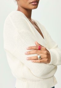 Hvid strikket cardigan med V-udskæring, der har knapdetaljer. Modellen har en rød ring og et skinnende sølvring på hånden.