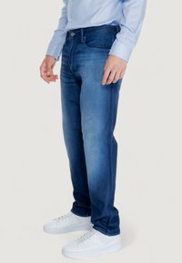 Jeans di denim blu scuro con una vestibilità slim e un leggero sbiadimento. L'outfit è completato da sneaker bianche e una camicia azzurra a maniche lunghe.