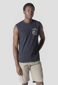 Joven de pie con expresión relajada, vistiendo una camiseta negra sin mangas con texto gráfico y pantalones cortos cargo beige.