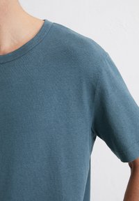 Grijs-blauwe katoen T-shirt met een ronde hals, korte mouwen en een geribbelde structuur. Netjes gestikte randen rond de halslijn en mouwen.