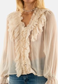 Blusa de gasa beige claro con volantes verticales en el frente, mangas largas con puños fruncidos y un corte relajado.