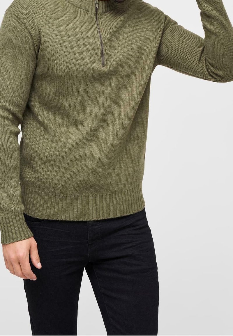 Homme portant un pull tricoté côtelé vert olive avec une fermeture éclair à l'avant et un pantalon noir, ajustant la manche sur fond blanc.