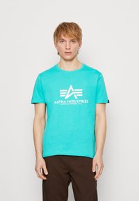 Türkisfarbenes Rundhals-T-Shirt aus Baumwolle mit weißem Logo-Druck und kurzen Ärmeln. Der Stoff hat eine glatte, leicht strukturierte Oberfläche.