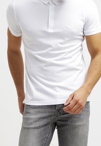 Vit bomullspolo med krage, korta ärmar och en treknappslåsd. Har en diskret logotyp på vänster bröst. Bärs med grå jeans.