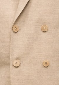 Μπεζ ύφασμα ψαροκόκκαλο με διπλό πέτο που διαθέτει τέσσερα ανοιχτό καφέ κουμπιά και μερικώς ορατό γιακά.