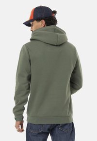 Haut à capuche vert avec une coupe décontractée et une grande capuche, présentant une texture douce et des poignets côtelés, porté avec un jean en denim.