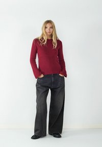 Roter gerippter Langarmpullover, tailliert, kombiniert mit weiten schwarzen Jeans und schwarzen Schuhen. Vor einem schlichten weißen Hintergrund.